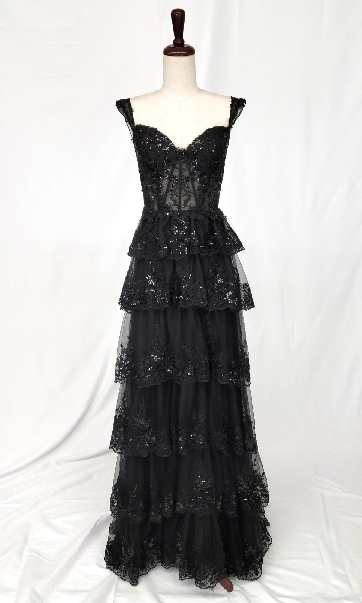 The Tiered Long Ruffle Prom Dress: Chicas C2433 is a black, floor-length, sleeveless sheer corset formal dress with a fitted bodice and tiered ruffled lace skirt, shown on a dress form against a plain white background.